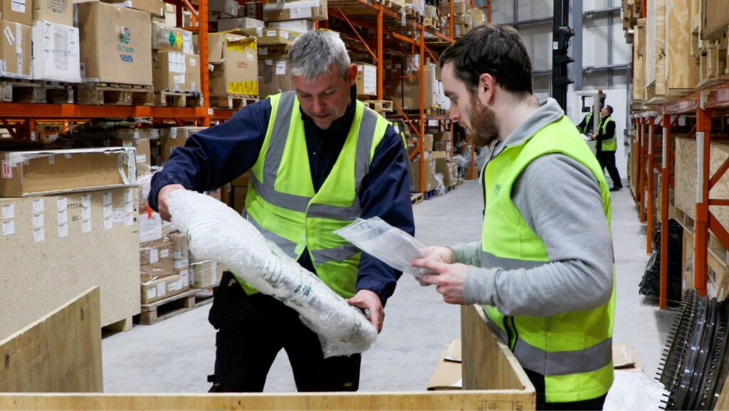 Two male EirTrade Aviation experts wearing safety jackets inspect a small aircraft component while standing in a warehouse. 