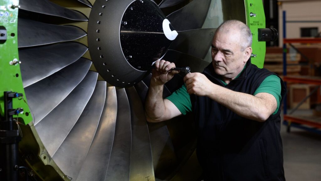 An aircraft engineer wearing a bodywarmer performs maintenance work on the turbine blades of a jet engine, with the workings partly exposed. 